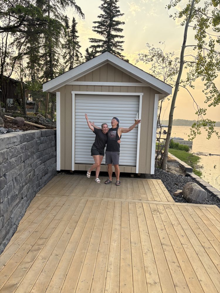 Two happy customers with arms raised in front of their new Apex shed on a cedar deck beside a lake