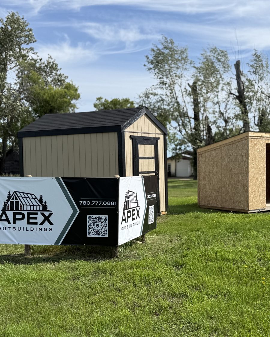 Three Apex sheds beside the Apex sign in the yard: a tan gable, one in bare OSB and a barn